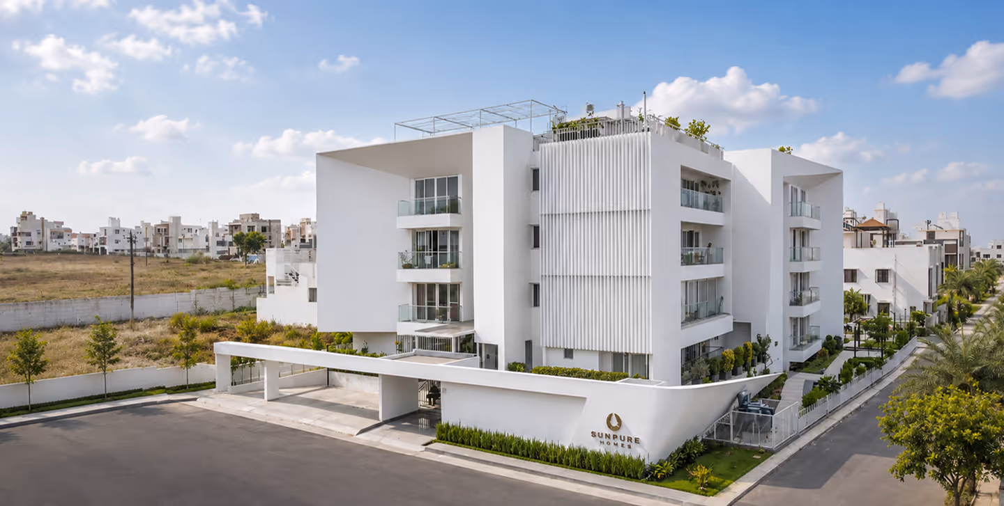Modern white residential building with balconies and greenery under a partly cloudy sky.