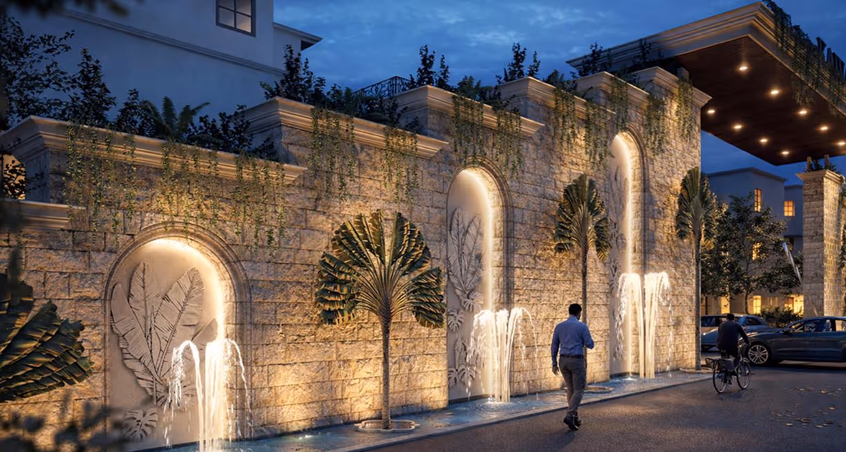 Evening view of a stone wall with lit water fountains and large leaf decorations along a driveway.