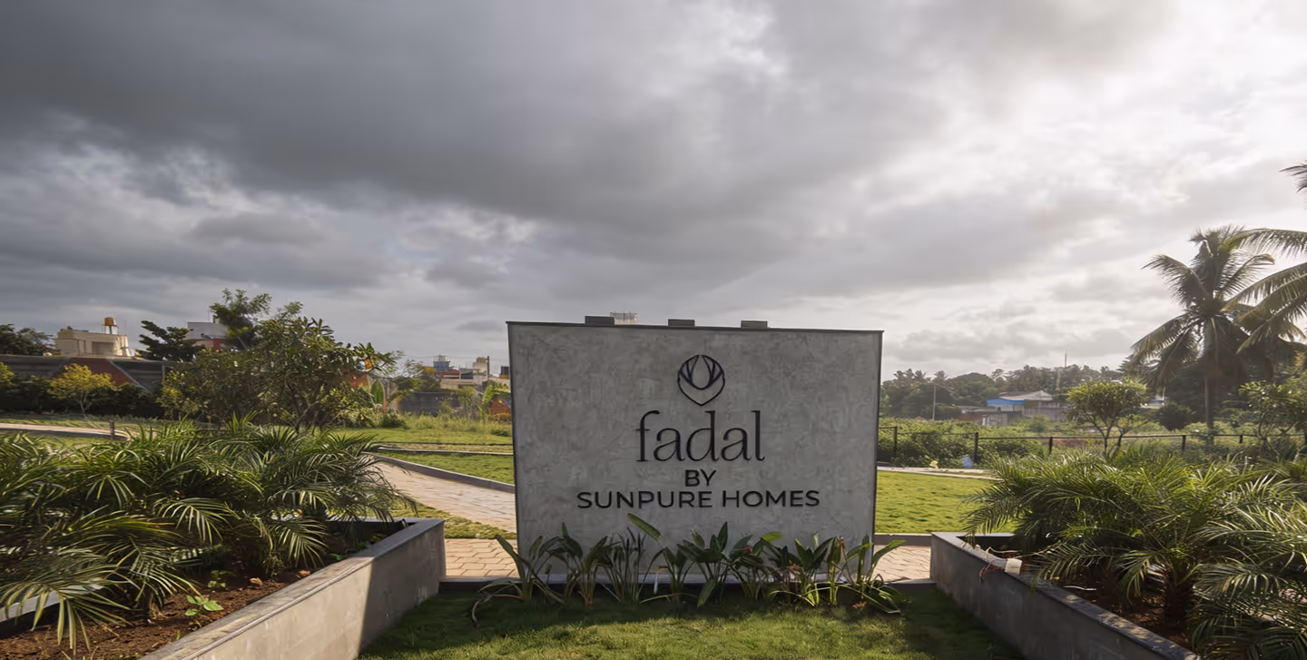 Concrete sign with text 'fadal BY SUNPURE HOMES' surrounded by greenery under a cloudy sky.