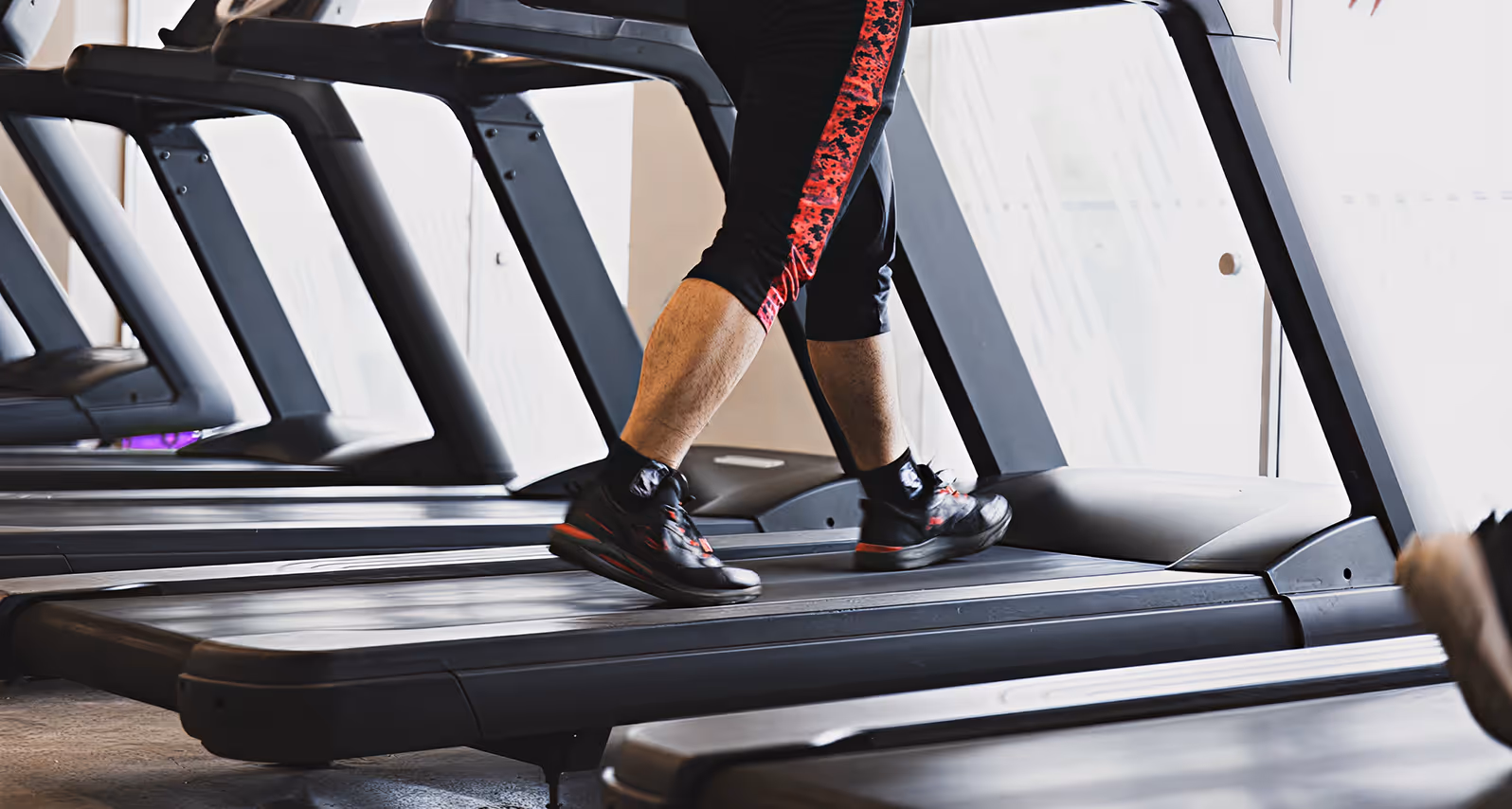 Person running on a treadmill wearing black shoes and black pants with red design in a gym.