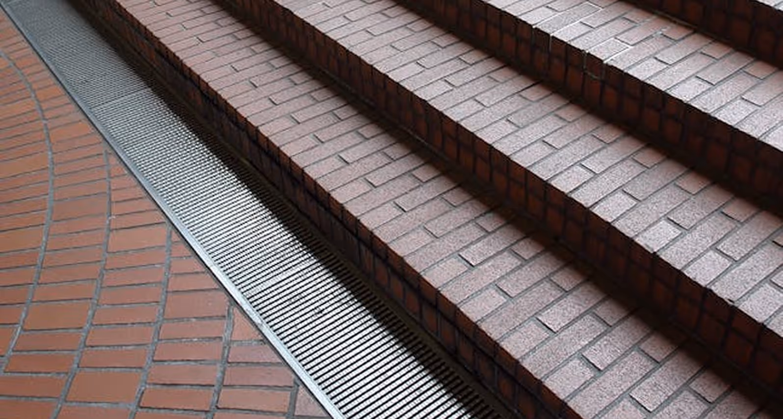 Brick stairs alongside a metal drainage grate embedded in a brick-tiled floor.