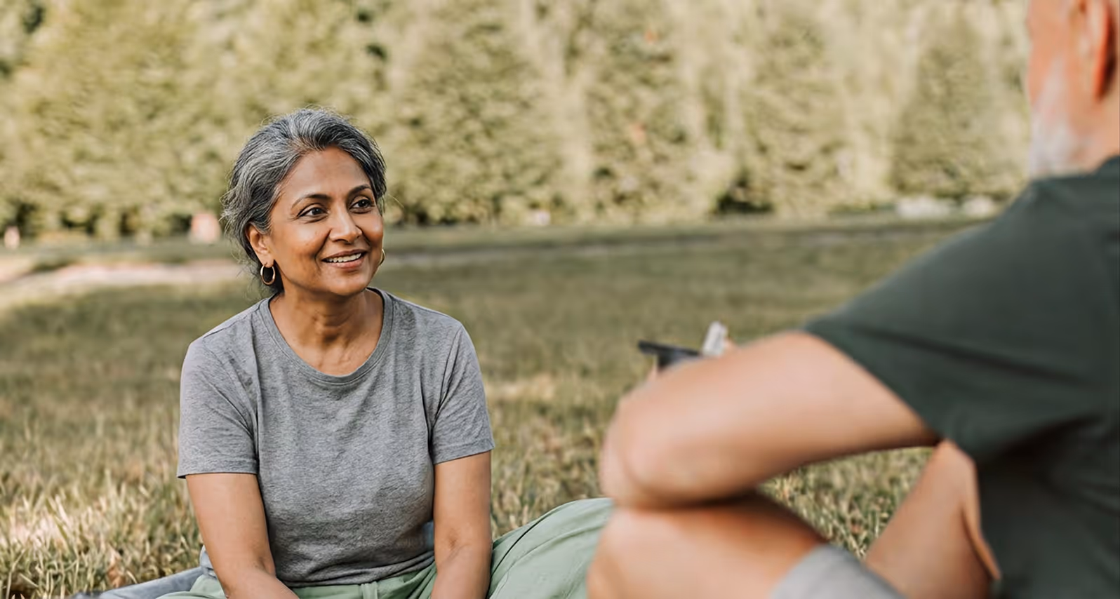 Smiling senior woman in gray shirt sitting on grass, talking to a person with folded arms in park.
