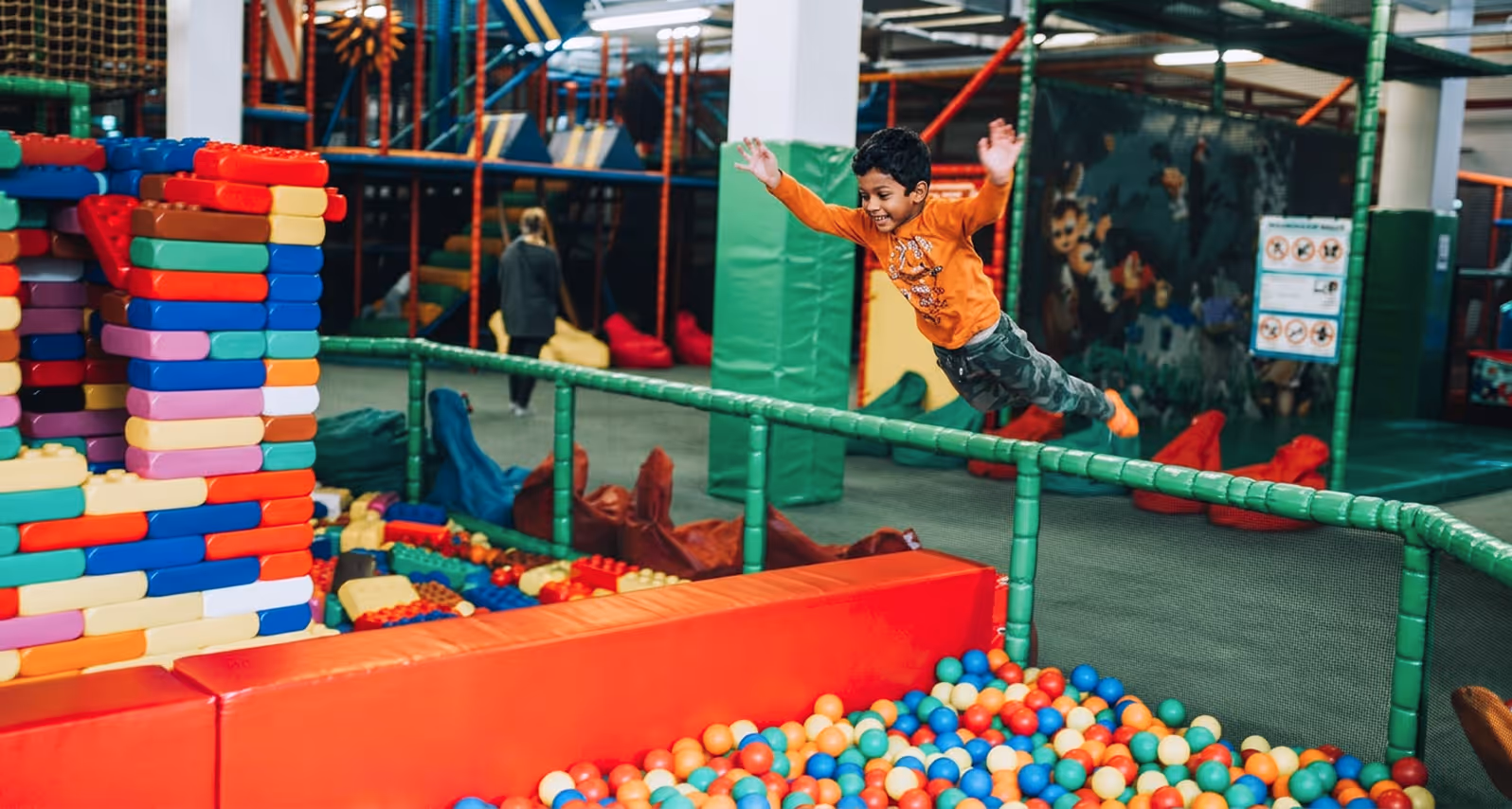 Child in orange shirt jumping into a ball pit in an indoor kids' play area with colorful blocks.