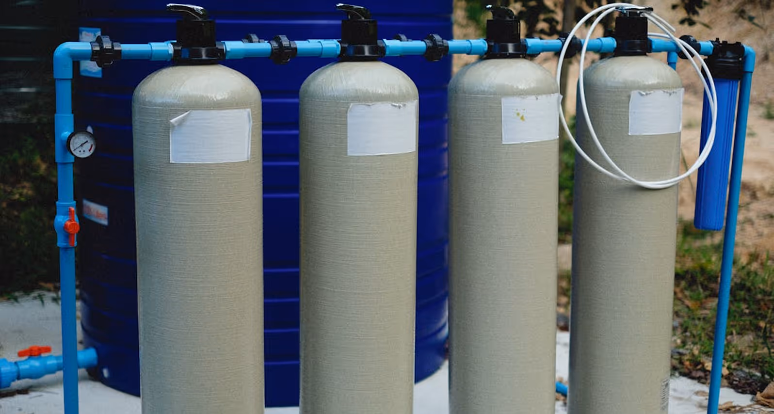 Four beige water filtration tanks connected by blue pipes with a pressure gauge and filter cartridge.