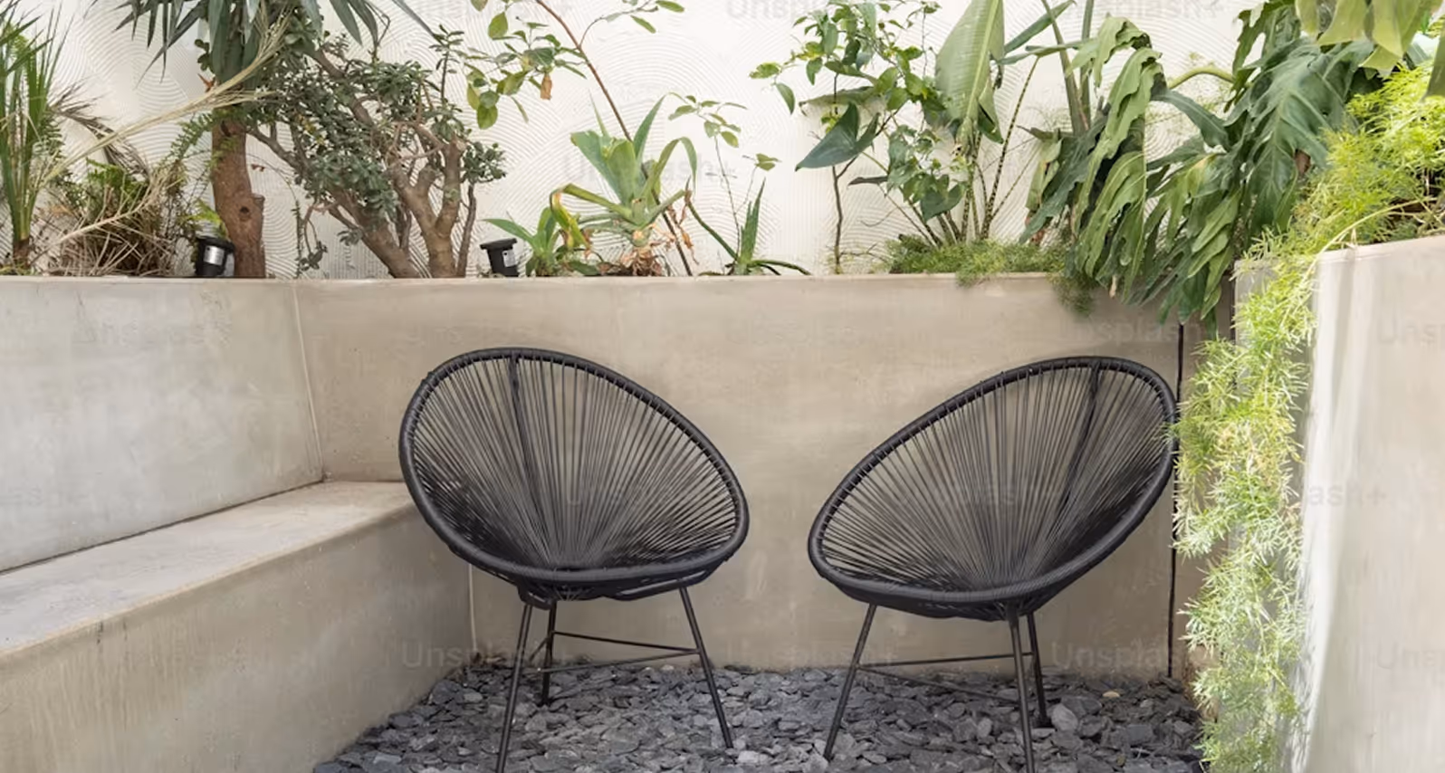 Two black woven chairs on gravel in a concrete corner with various green potted plants behind.