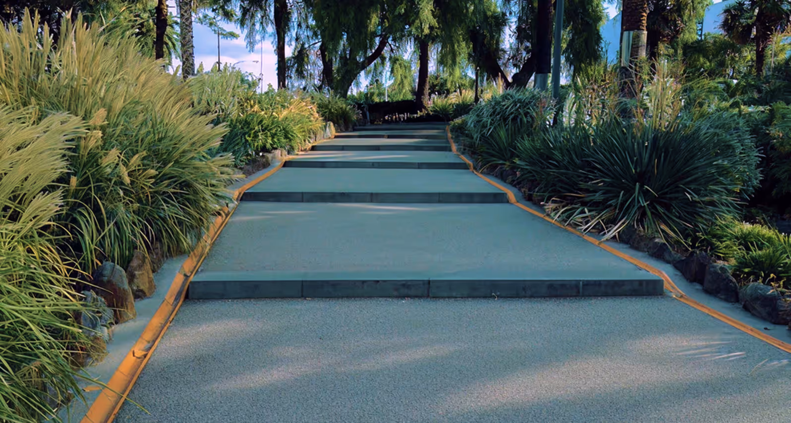 Concrete pathway with small steps bordered by lush green plants and trees in a landscaped garden.