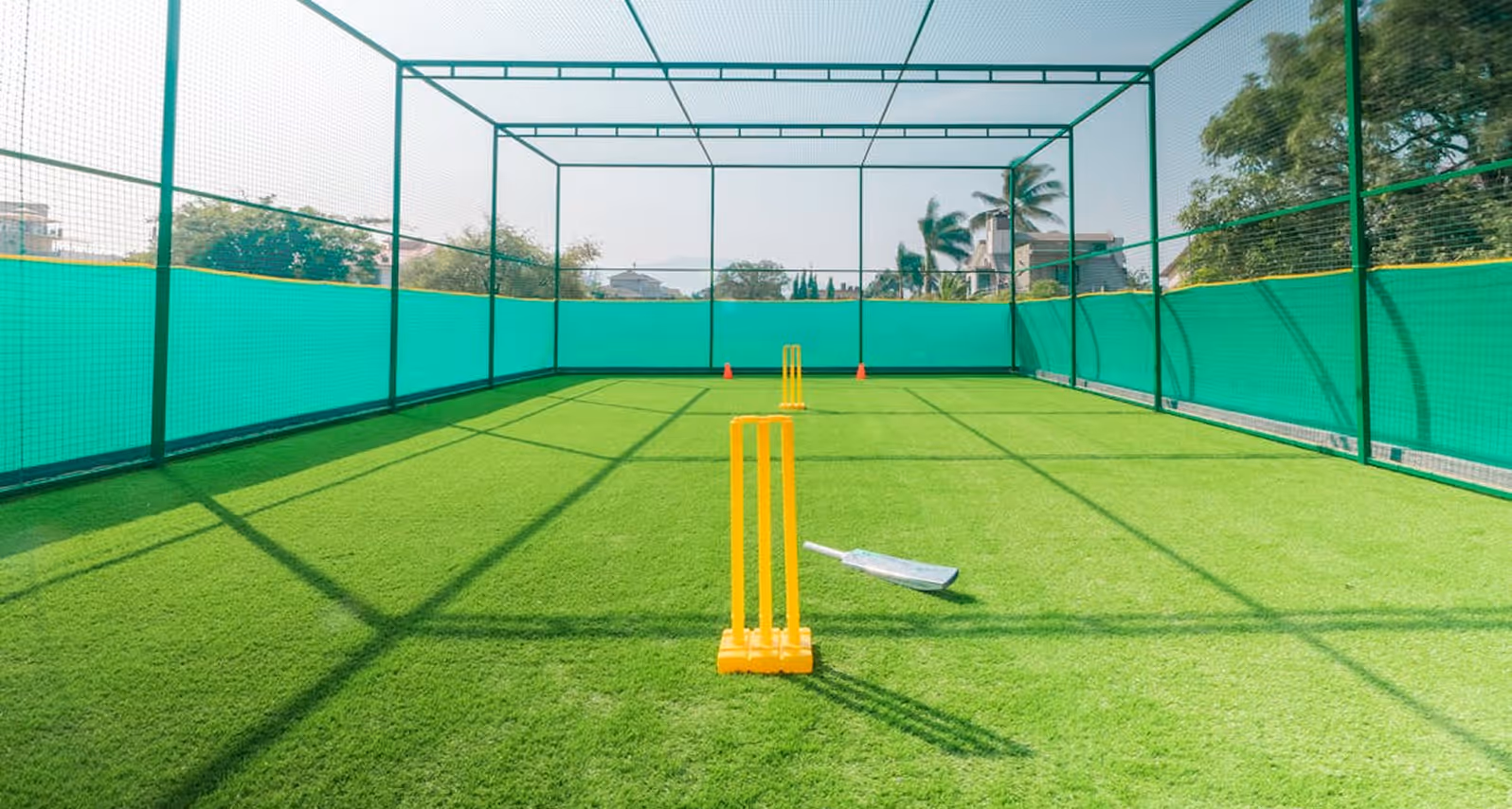 Outdoor cricket practice net with green turf, yellow stumps, and a cricket bat lying on the ground.