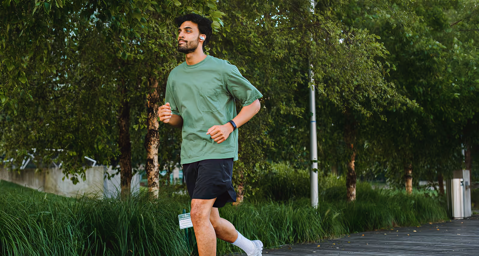 Man jogging on a wooden path surrounded by lush green trees and grass wearing headphones and fitness tracker.