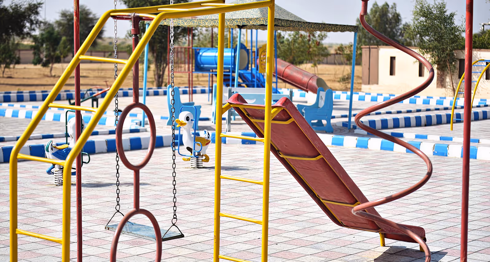 Children's playground with slide, swings, spring riders, climbing structures, and a shaded bench area.