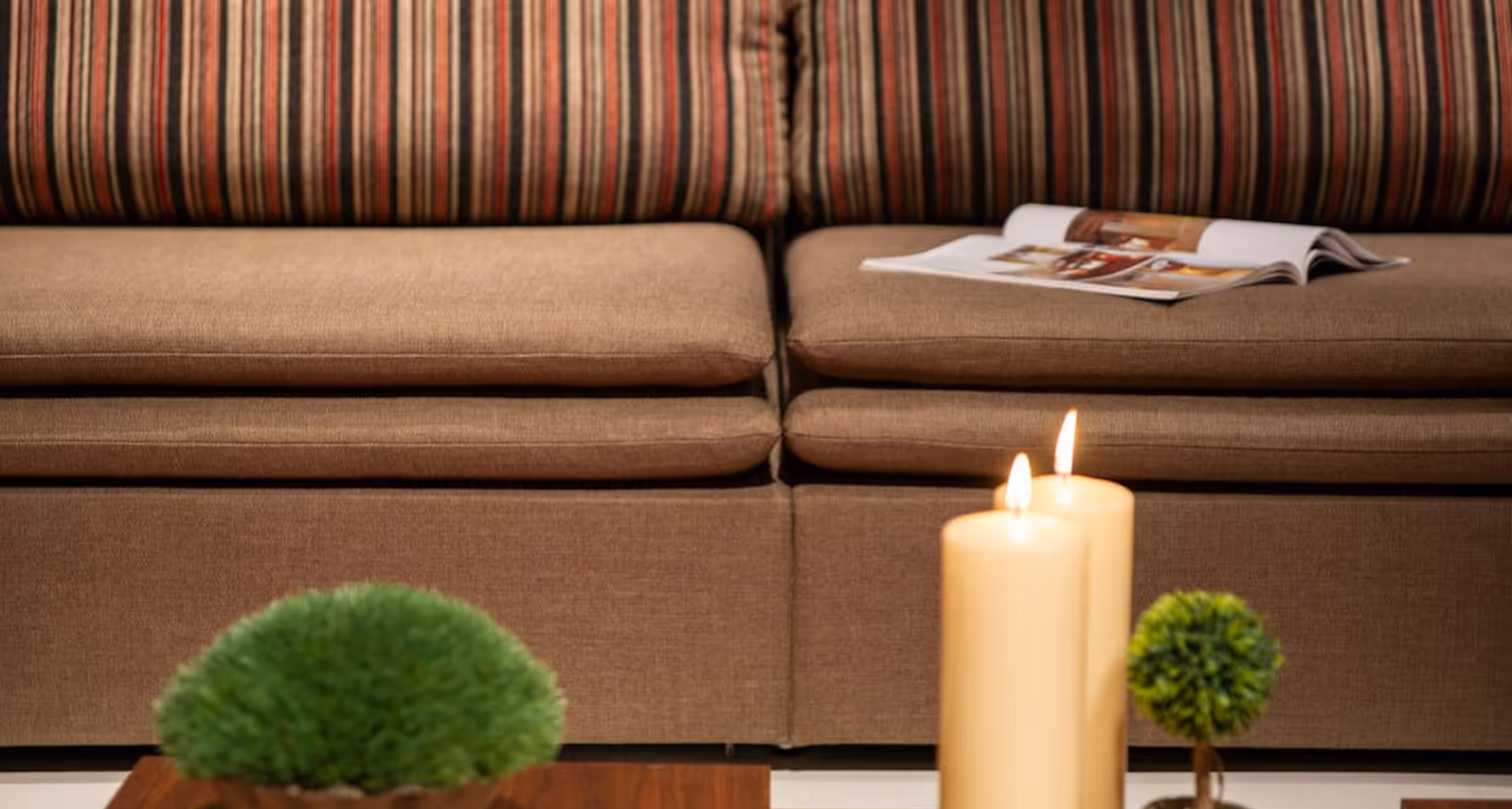 Brown sofa with striped backrest, magazine, two lit candles, and small green plants on a wooden table.