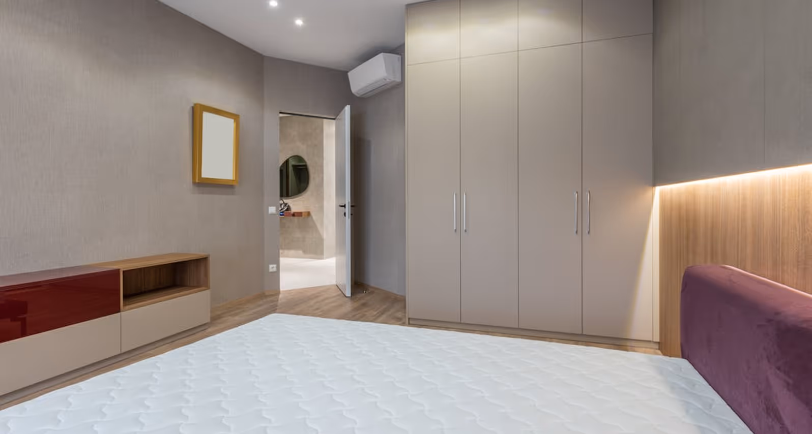 Minimalist bedroom with white bed, purple headboard, beige wardrobe, and open door to bathroom.