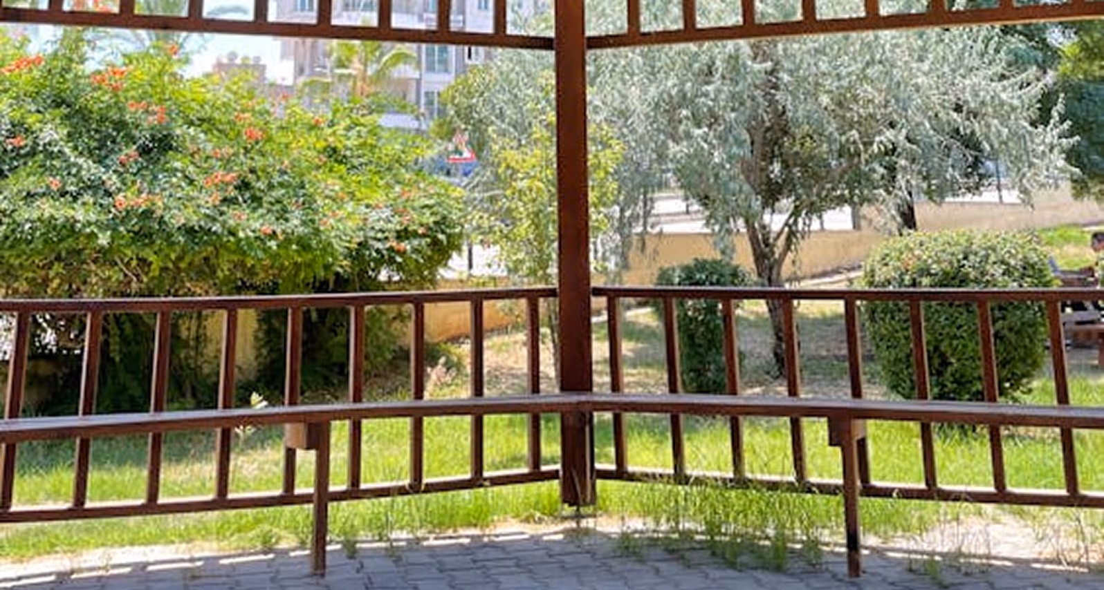 Empty wooden bench seating under a pavilion with green grass, bushes, and flowers outside.
