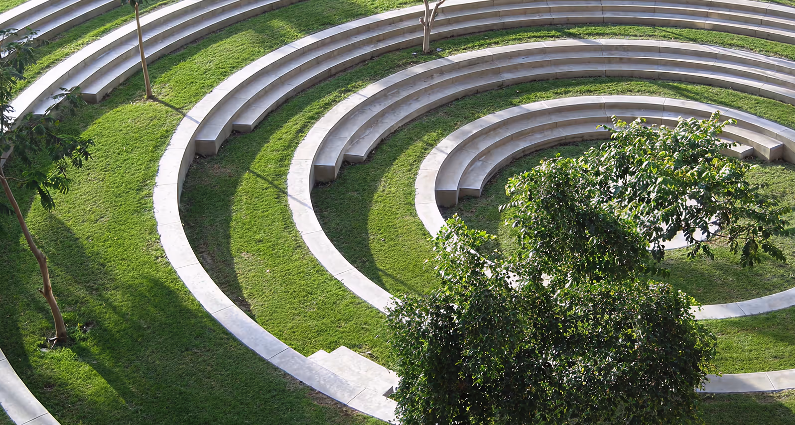 Outdoor amphitheater with curved concrete seating surrounded by green grass and trees.