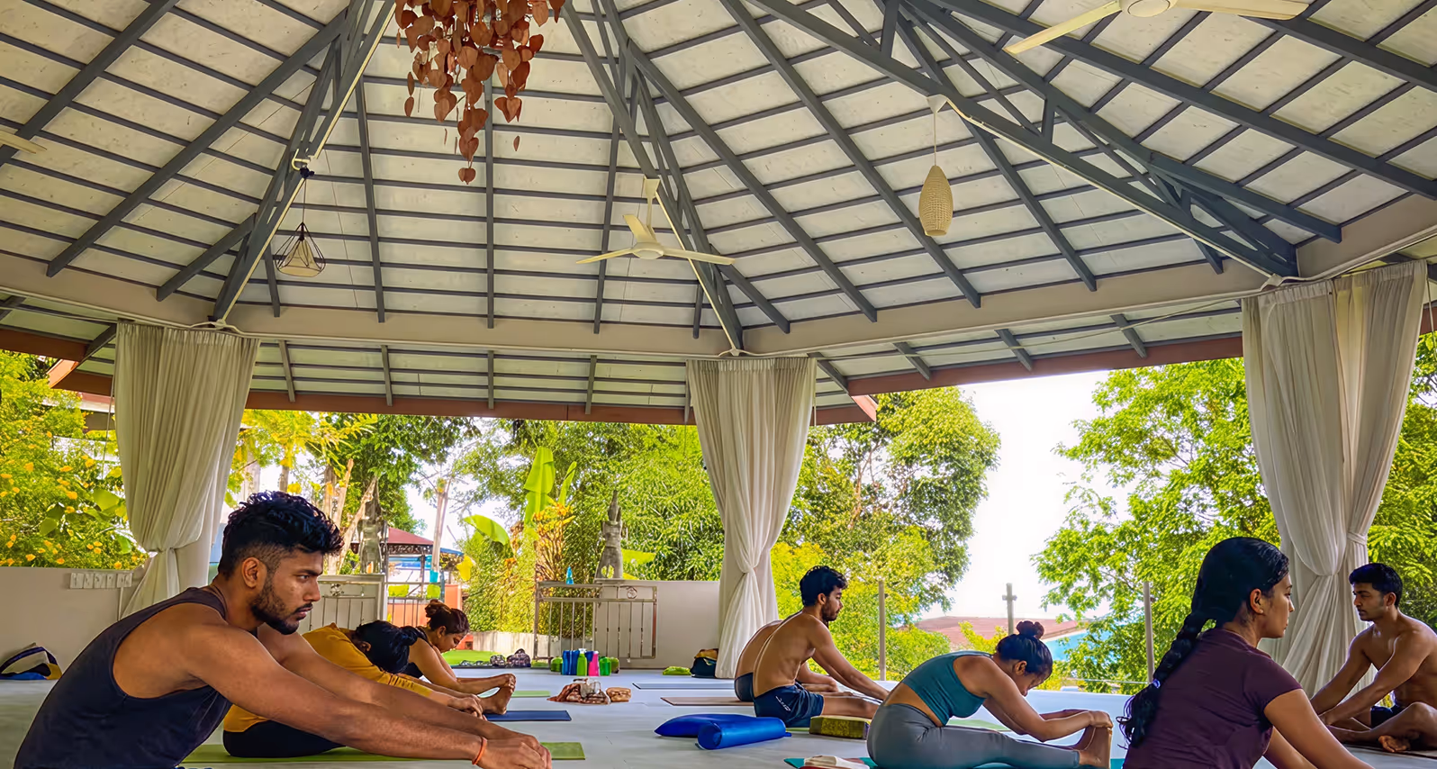 Group of people stretching indoors in a partially open yoga pavilion surrounded by greenery
