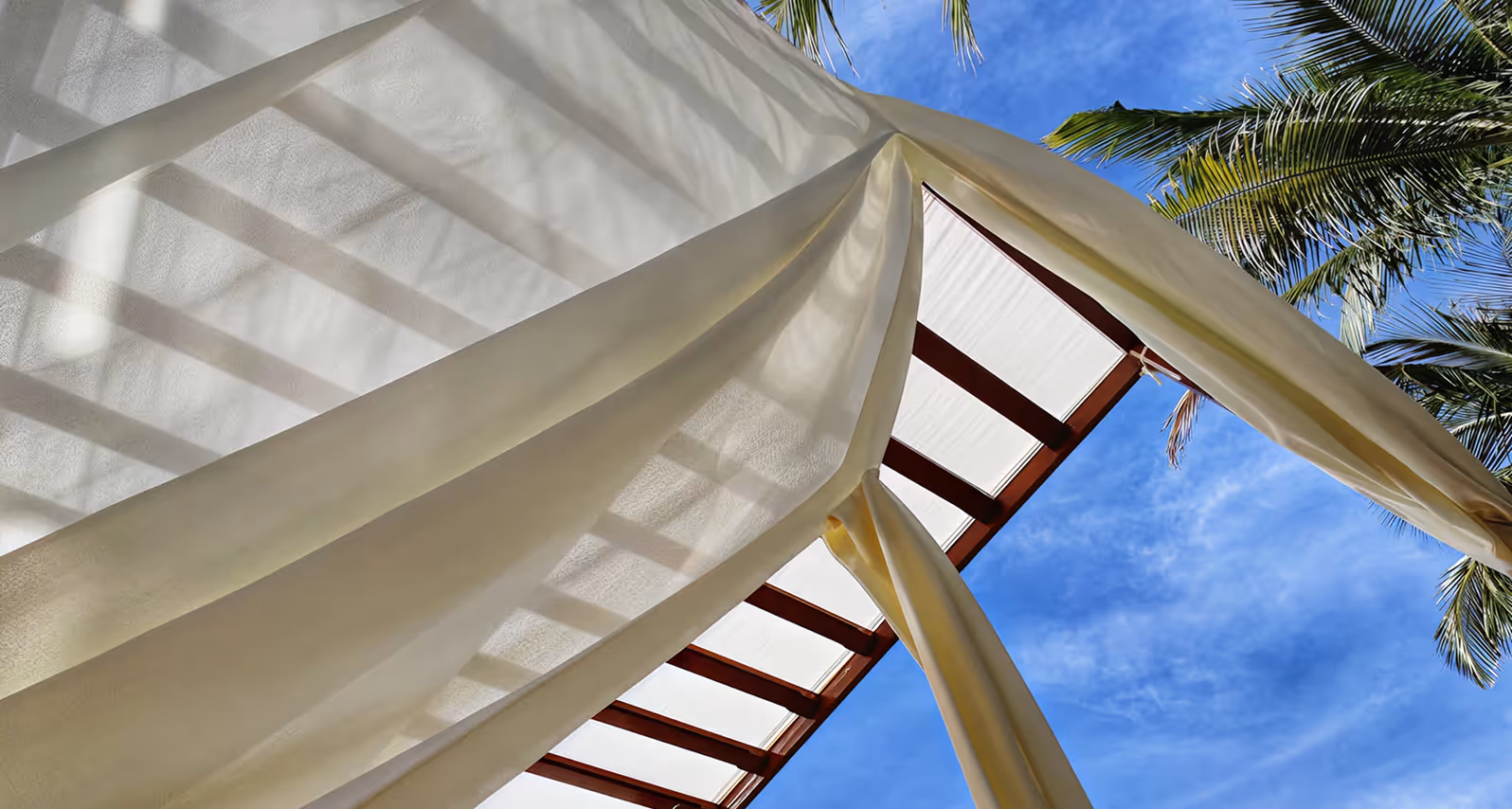 White curtains tied back on a pavilion with palm trees and a blue sky in the background.