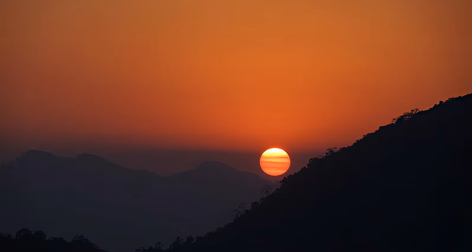 Sun setting behind a dark mountain silhouette with an orange sky fading to darker shades.