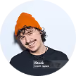 Mann mit lockigem Haar und orangefarbener Mütze lächelt vor hellem Hintergrund.