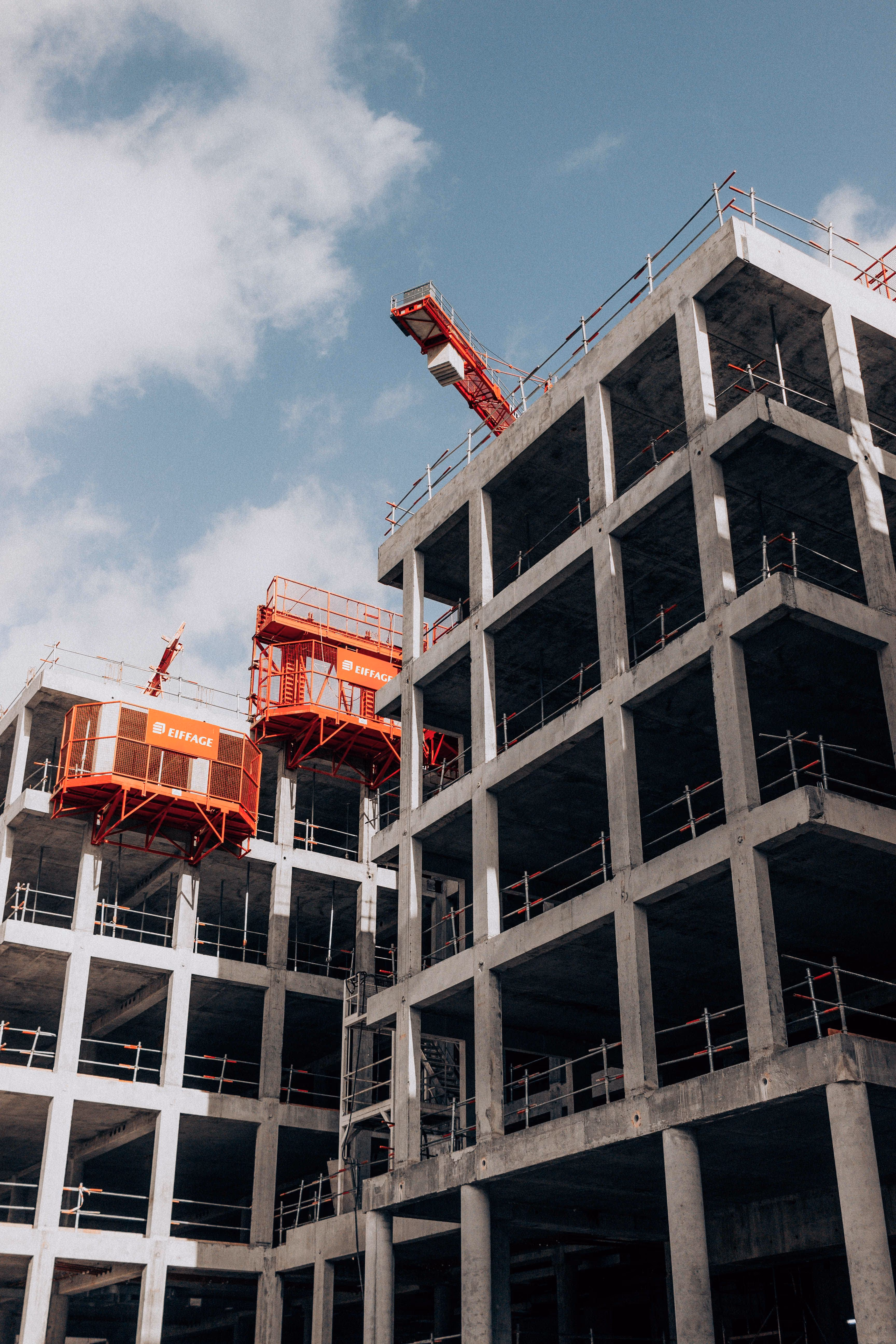 Unfertiges Betongebäude mit roter Bauaufzugs- und Kranstruktur vor blauem Himmel mit Wolken.