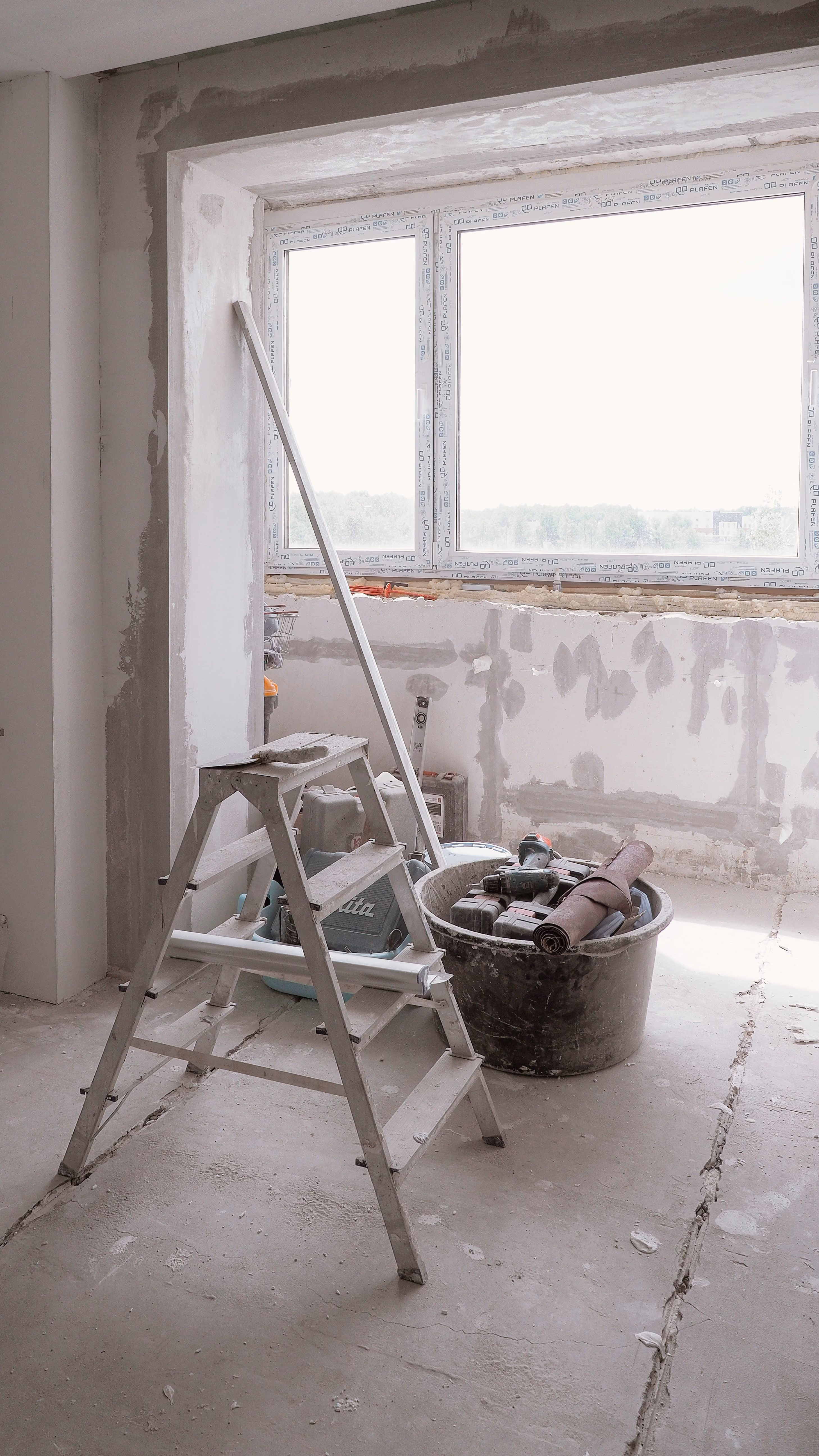 Baustelle eines Innenraums mit ungeputzten Wänden und einem großen Fenster, davor eine Stehleiter und ein Eimer mit Werkzeugen und Baumaterialien.