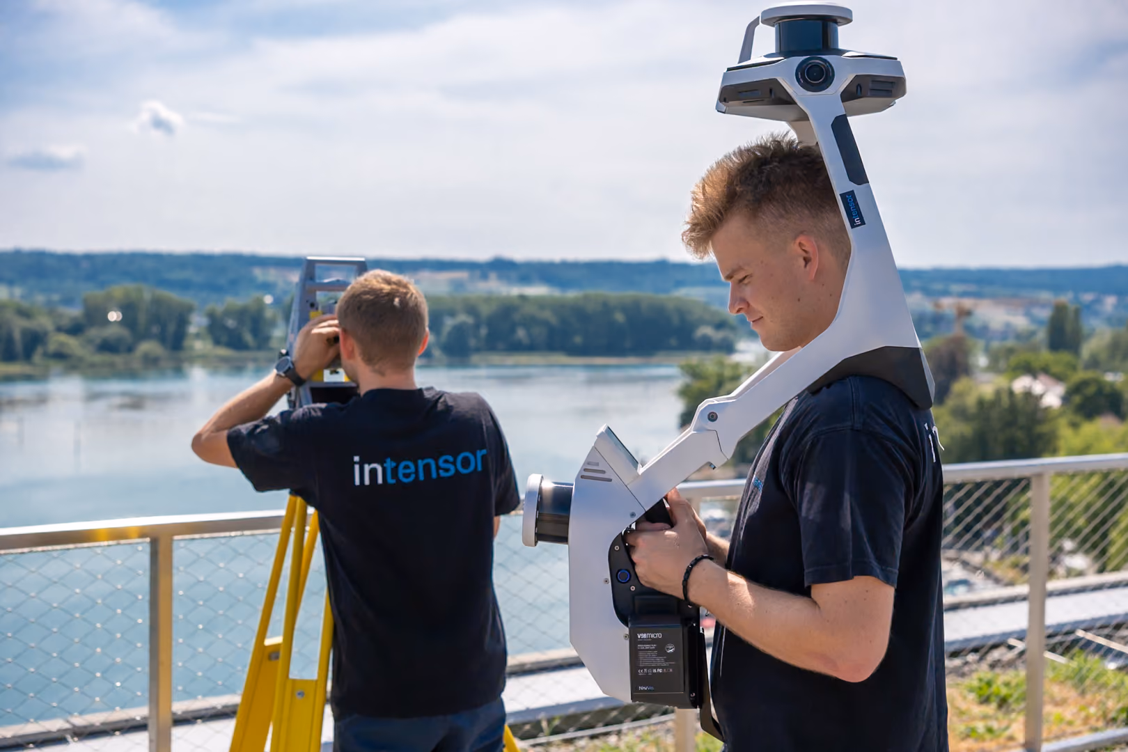 Zwei Vermessungsingenieure mit Vermessungsgeräten vor Fluss und Landschaft im Hintergrund.