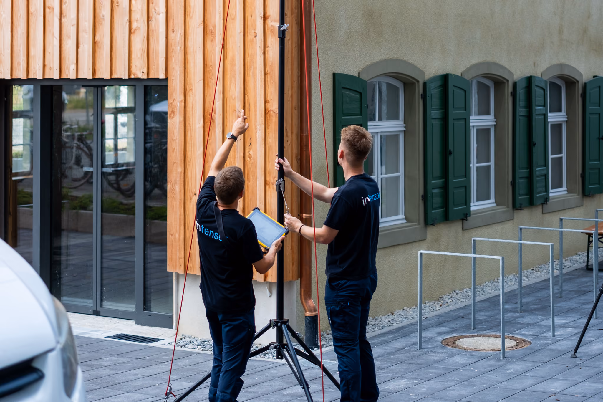 Zwei Männer in schwarzer Arbeitskleidung prüfen eine Stange vor einem Holz- und Fenstergebäude.