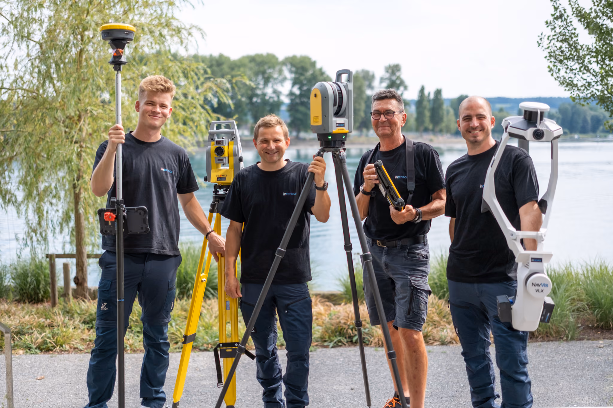 Vier Männer in schwarzer Arbeitskleidung stehen draußen auf einem Weg vor einem See, jeder hält verschiedene professionelle Vermessungsgeräte wie ein GPS-Empfänger, ein Tachymeter auf einem Stativ, ein Lasergerät und ein mobiles 3D-Laserscanning-System.