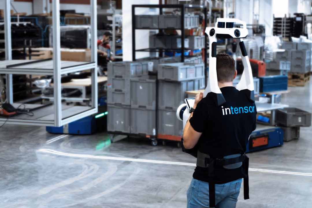 Mann mit schwarzem Intensorn-Shirt bedient ein mobiles 3D-Scan-Gerät in einer Industriehalle, umgeben von Regalen, Kisten und Ausrüstungen.