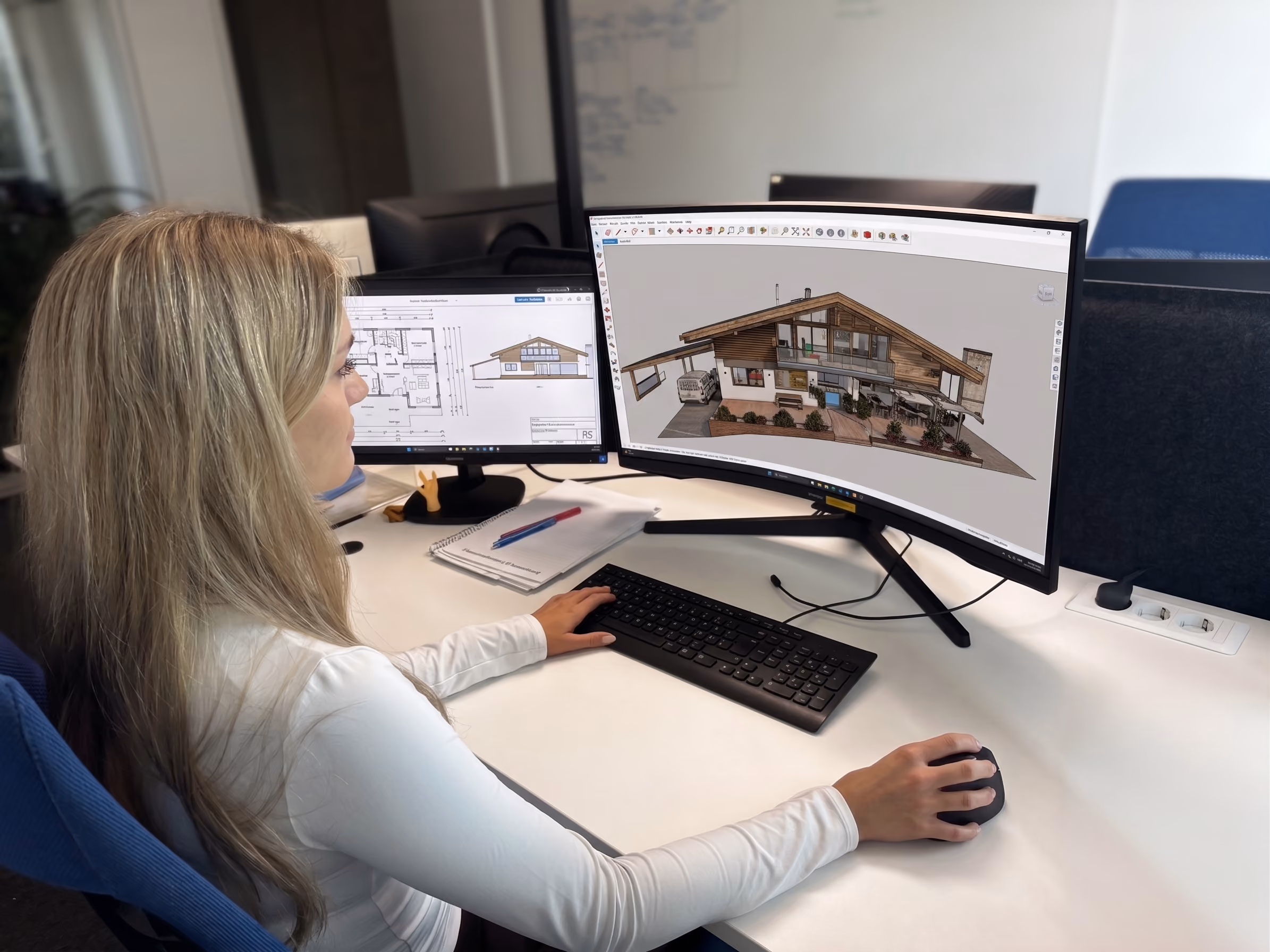 Frau sitzt an einem Schreibtisch vor zwei Bildschirmen, auf denen Architekturpläne und eine 3D-Visualisierung eines Hauses gezeigt werden.