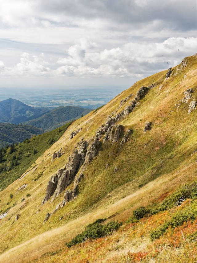 Sanfter Berghang mit Gras, Felsen und bewachsenen Hügeln unter bewölktem Himmel.