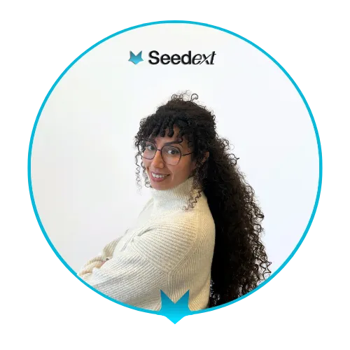 Portrait souriant d'une femme aux cheveux bouclés longs, portant un pull blanc et des lunettes, sur fond blanc avec le logo Seedext.