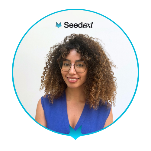 Portrait souriant d'une femme aux cheveux bouclés longs, portant un pull blanc et des lunettes, sur fond blanc avec le logo Seedext.