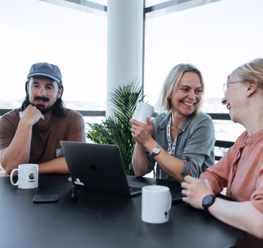 A group of four colleagues sitting around a table with laptops and coffee cups, smiling and having a discussion in a bright office space.