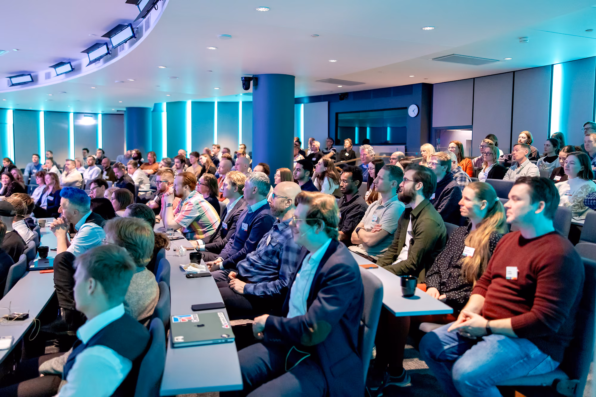 Audience attentively listening in a modern conference room with blue lighting and tiered seating.