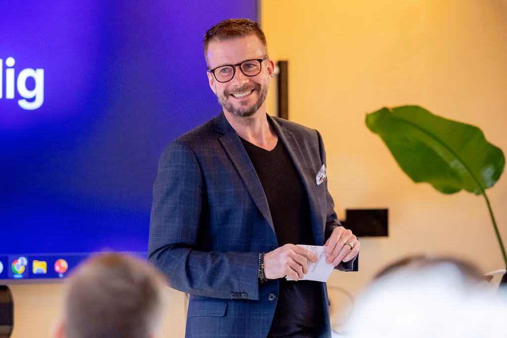 Smiling man in glasses and blazer speaking to an audience with a screen behind him.