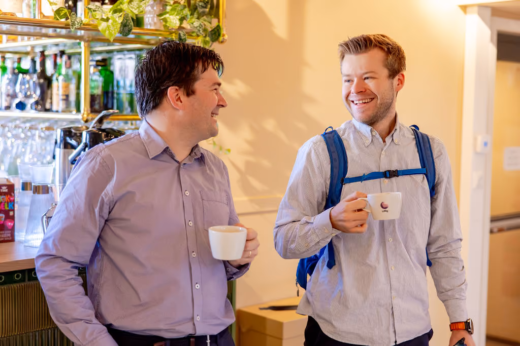 Two men smiling and holding coffee cups, one with a backpack strap across his chest.