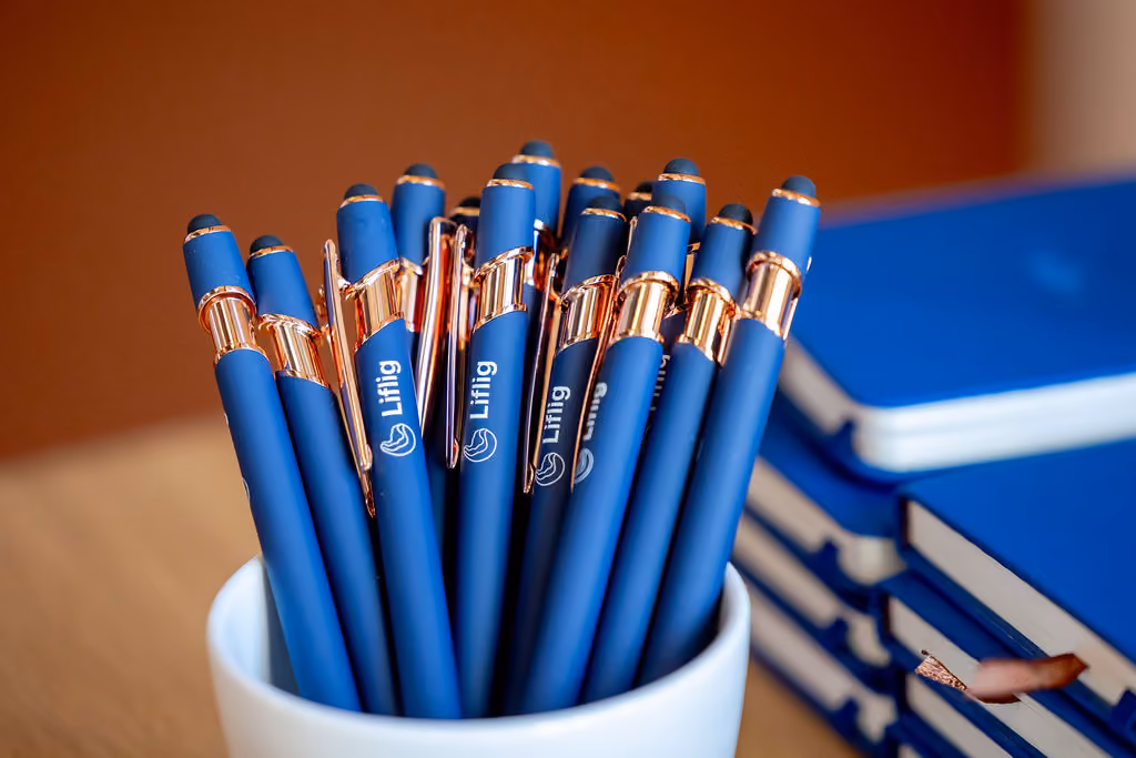 Blue pens with gold clips in a white cup next to stacked blue notebooks on a wooden table.