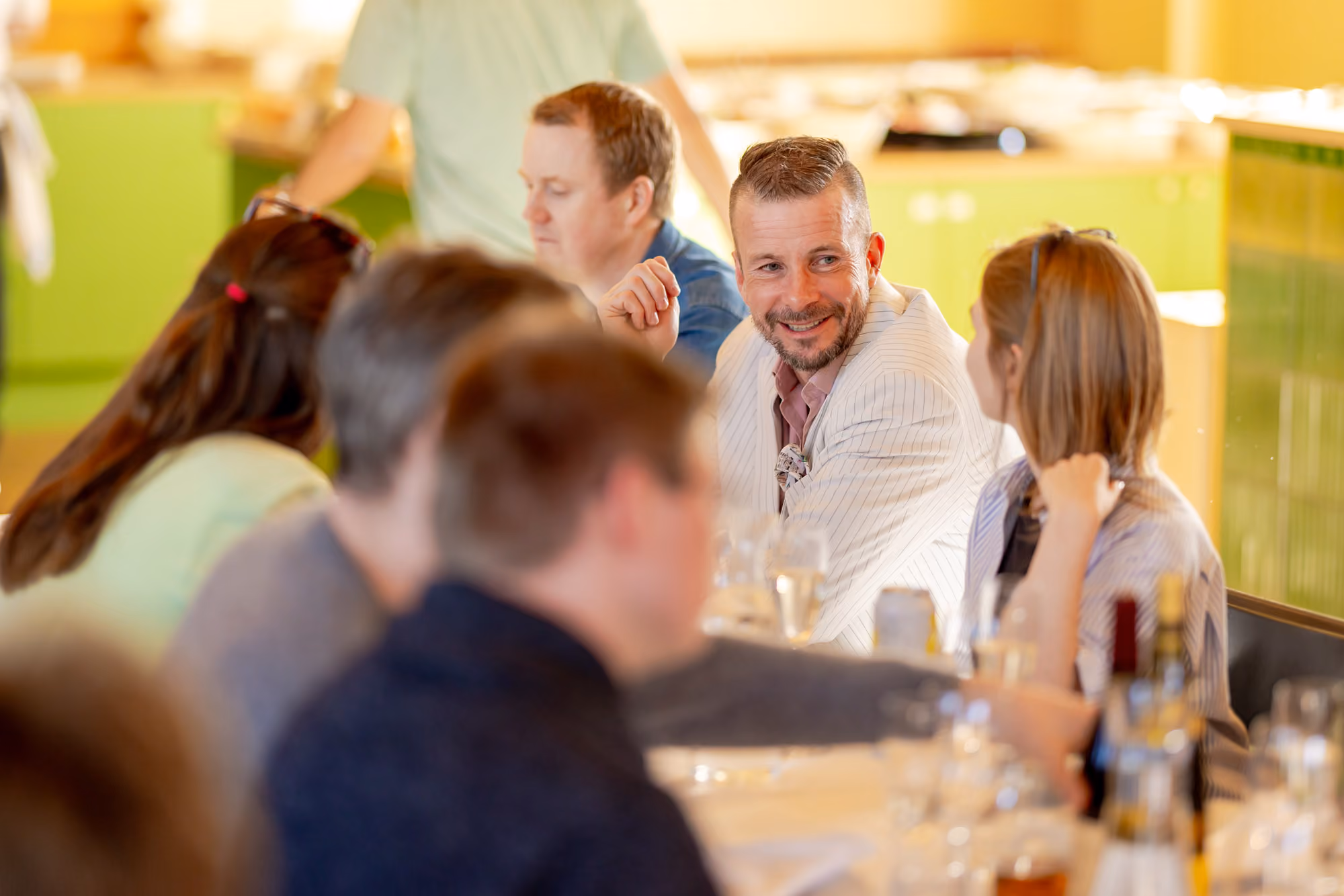A man in a white jacket smiles while talking to a woman at a table with drinks in a bright room.
