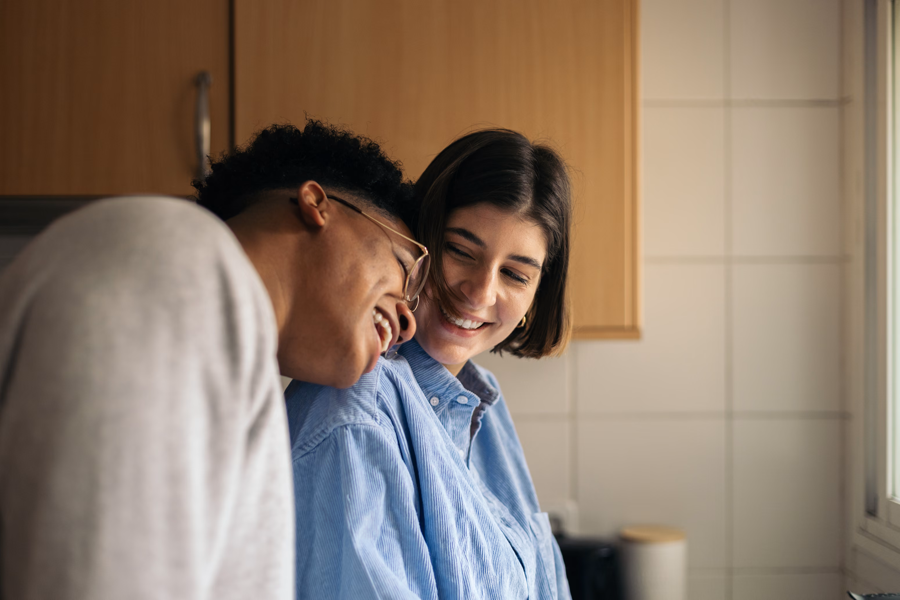 A man laughs leaning on a smiling woman's shoulder in a warmly lit kitchen.