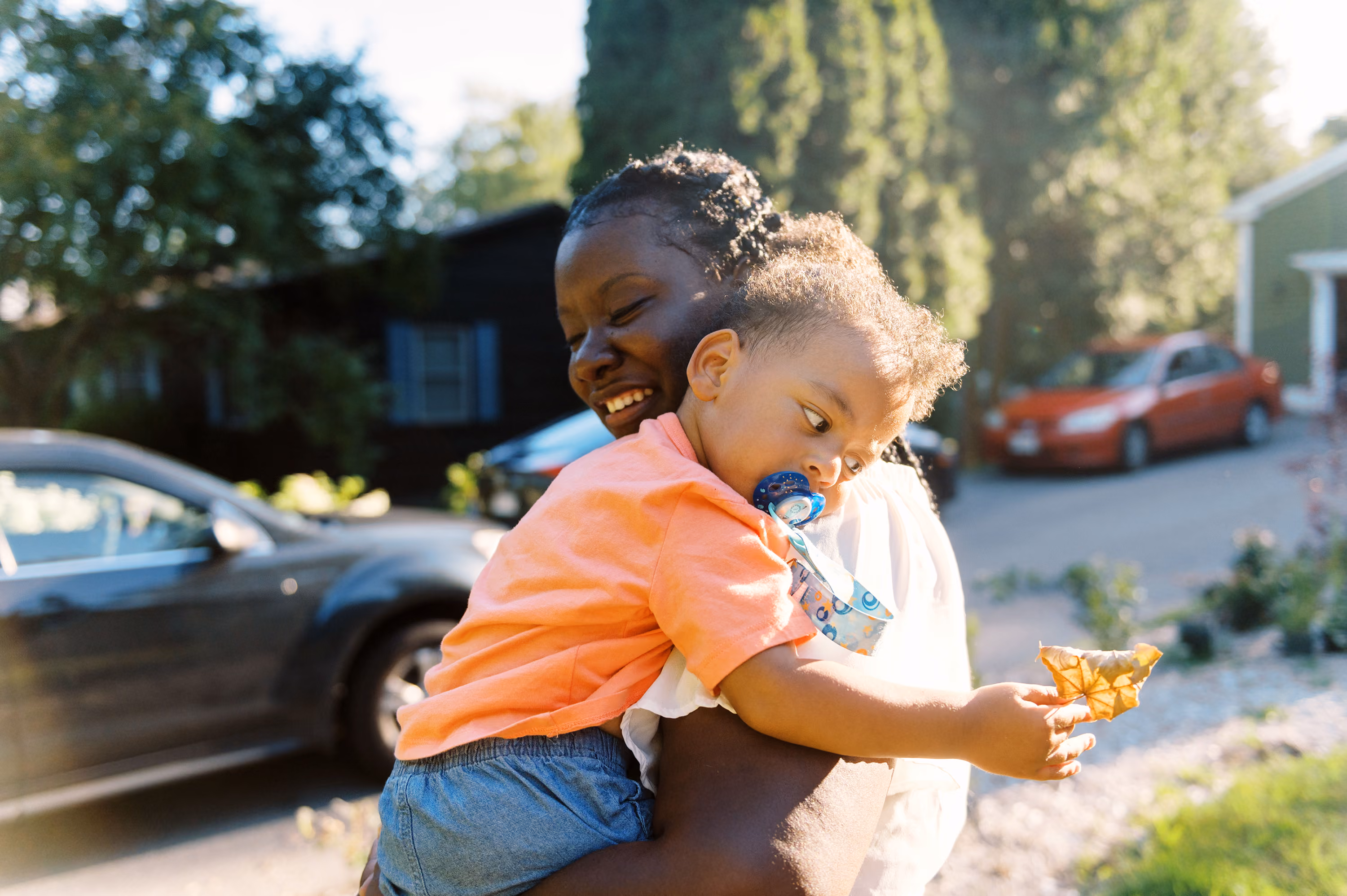 Smiling woman holding her child with a pacifier who is examining a leaf outdoors.