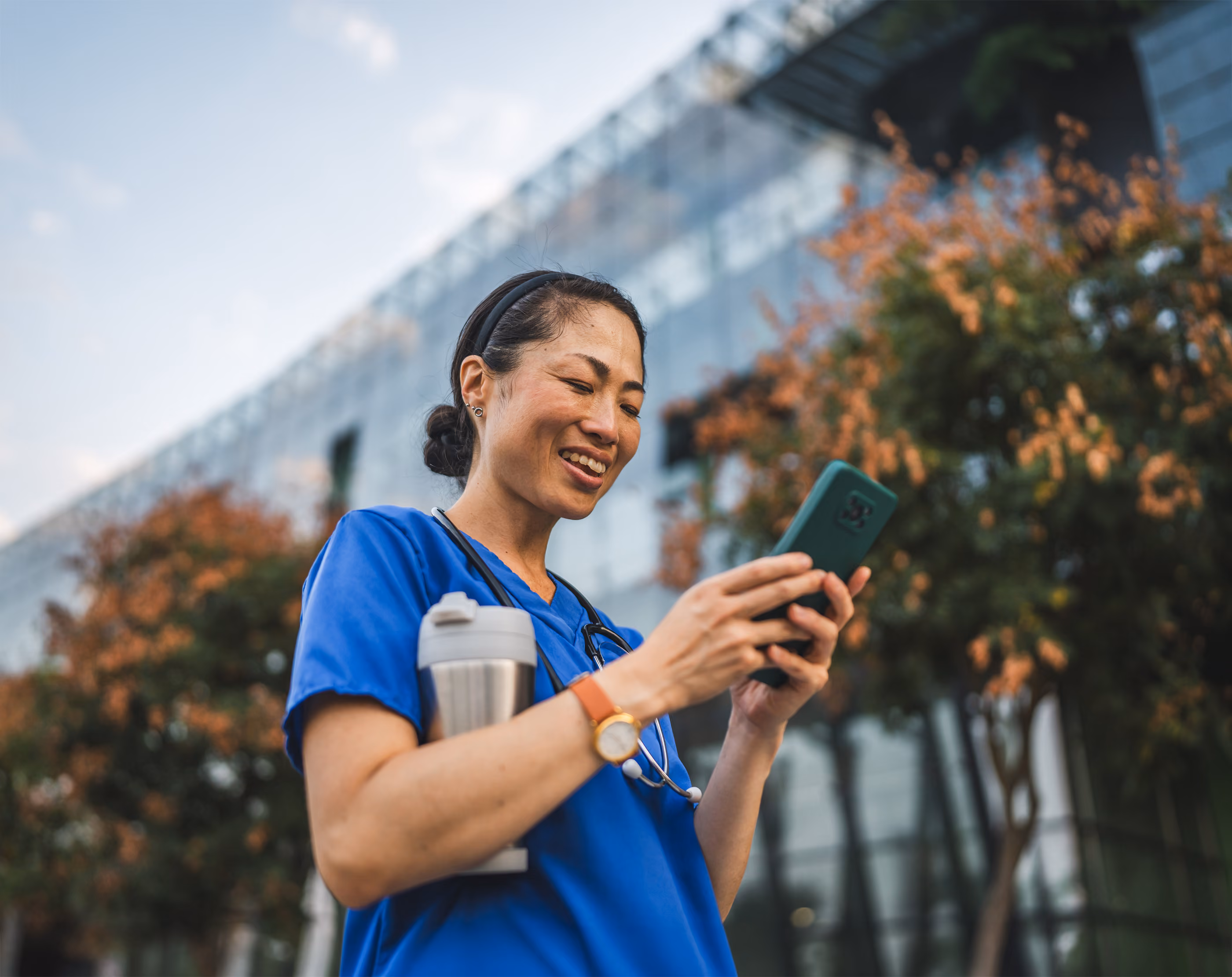 Smiling nurse in blue scrubs holding a coffee mug and using a smartphone outside a modern building.