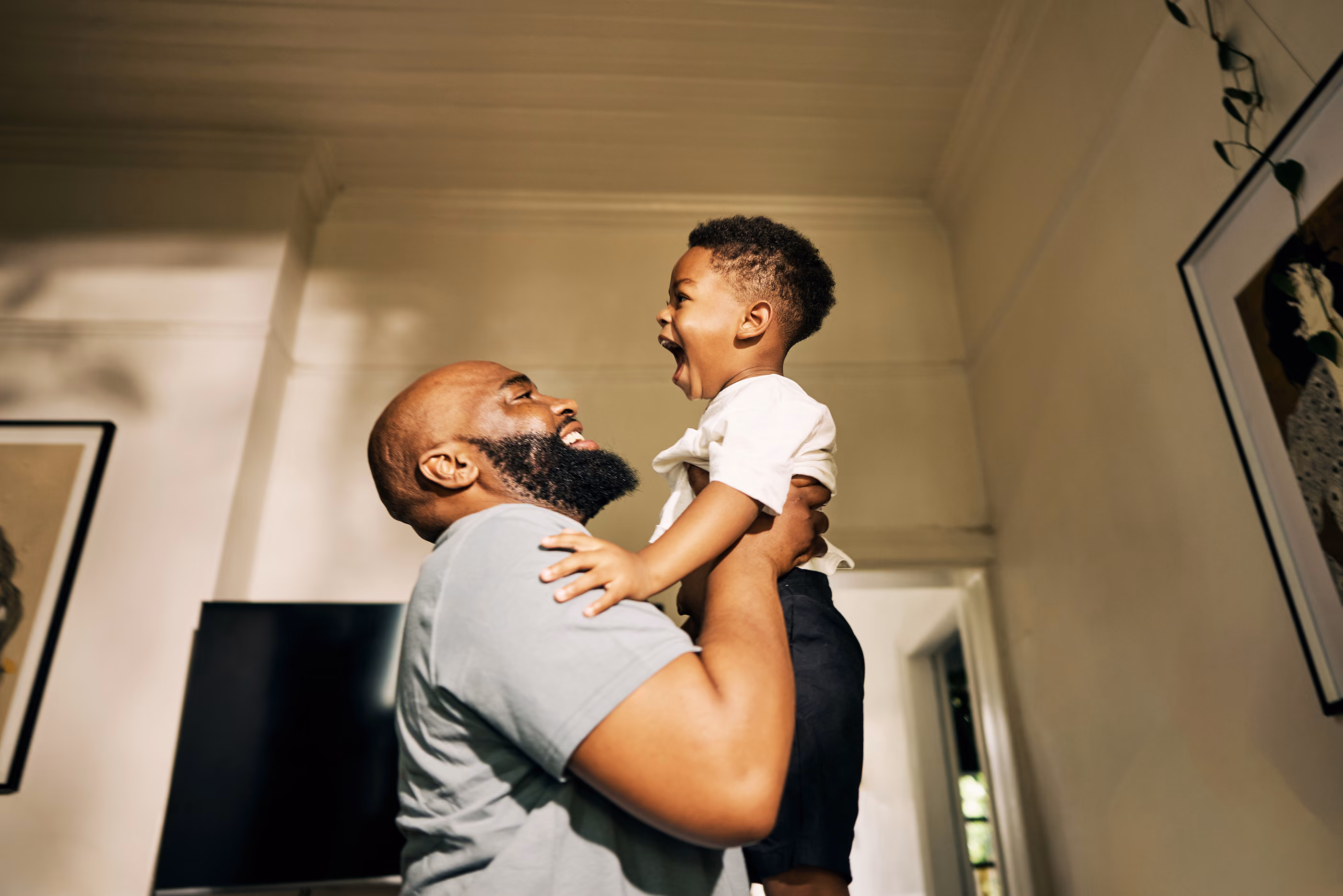 Man lifting smiling boy, both looking at each other joyfully indoors.