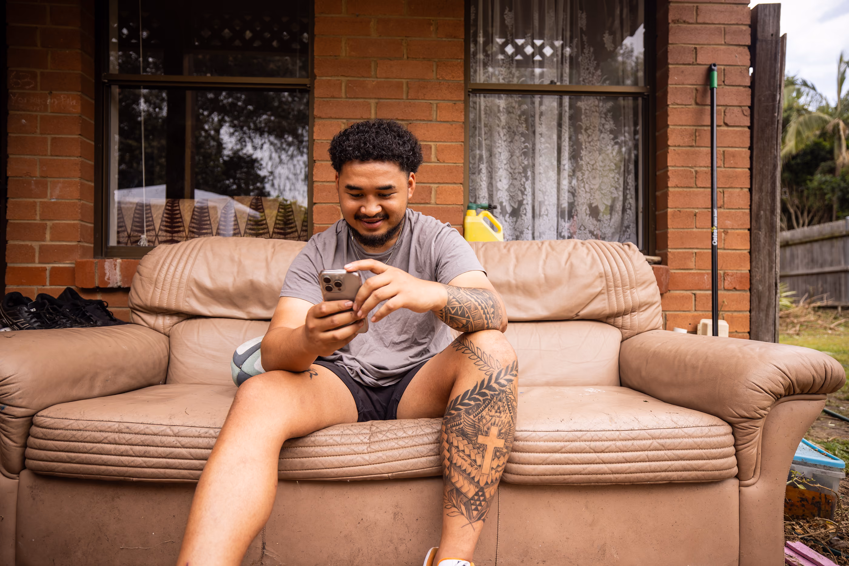 Man with tattoos on leg and arm sitting on worn couch outside, smiling while using smartphone.