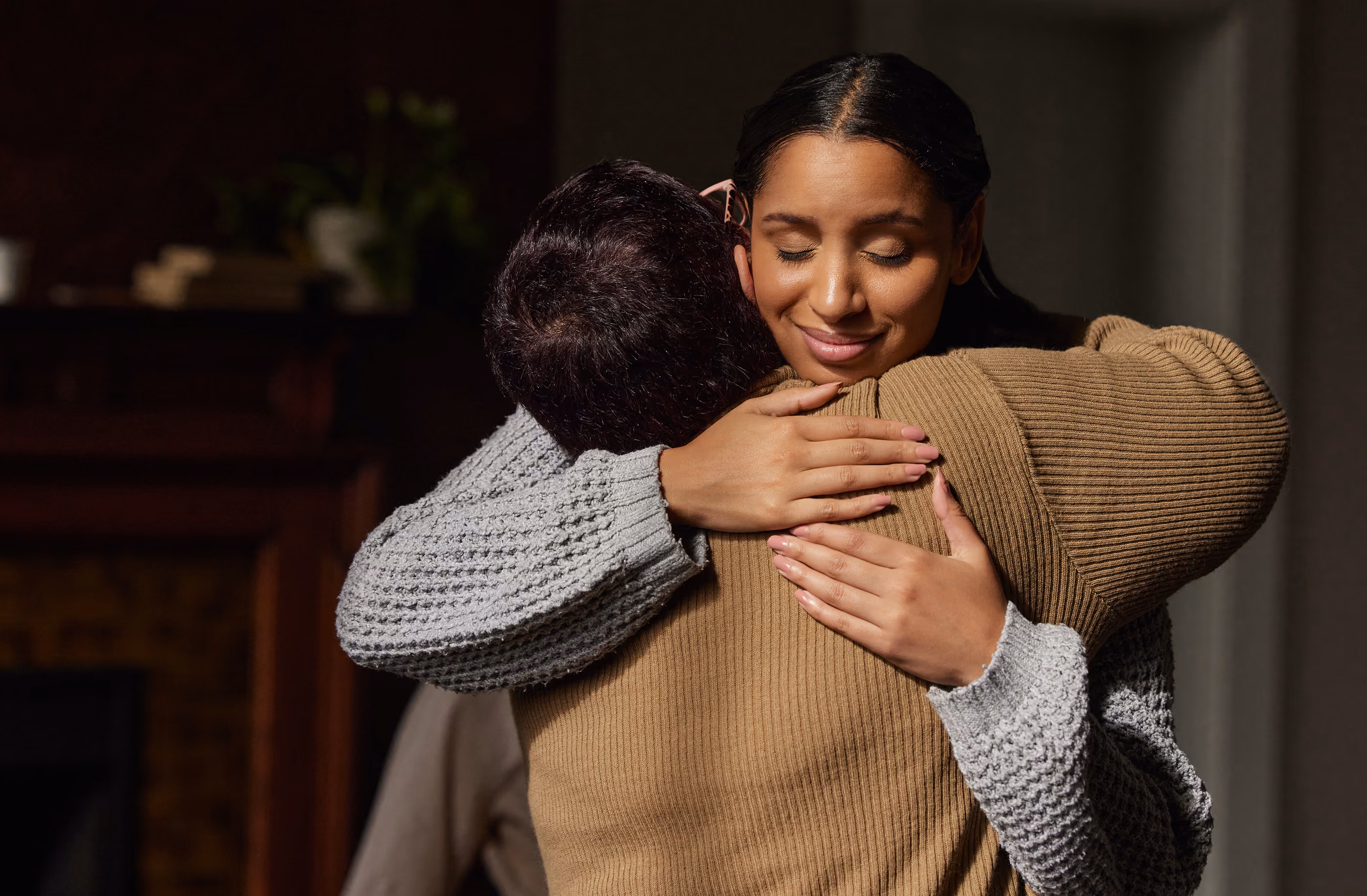Two people embracing tightly in a warm, heartfelt hug indoors with closed eyes.