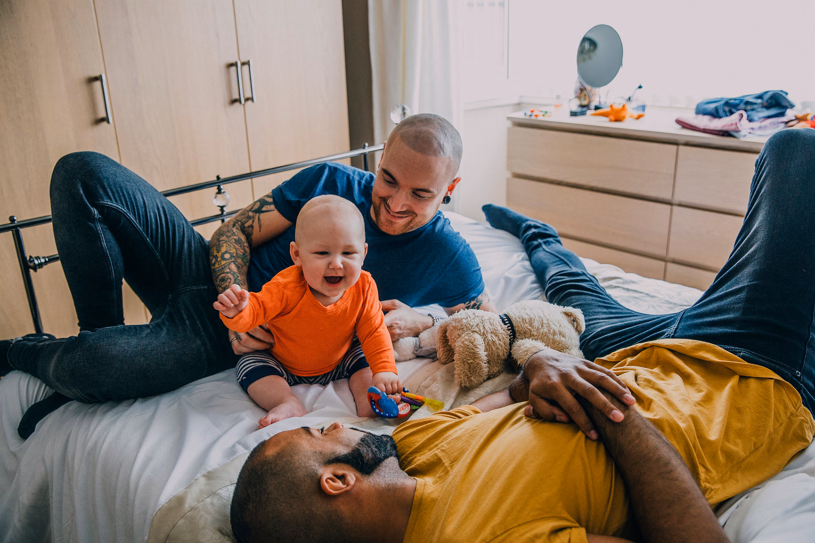 Two men and a baby playing happily on a bed with a stuffed dog nearby.