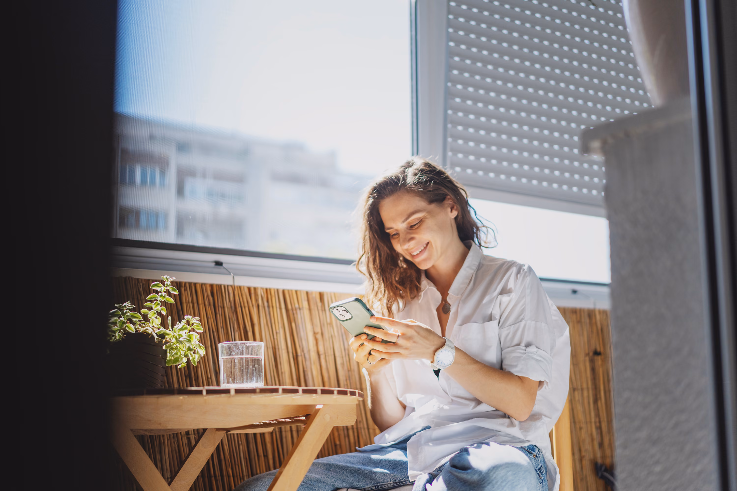 Smiling woman in white shirt sitting by a wooden table, looking at her phone near a window.