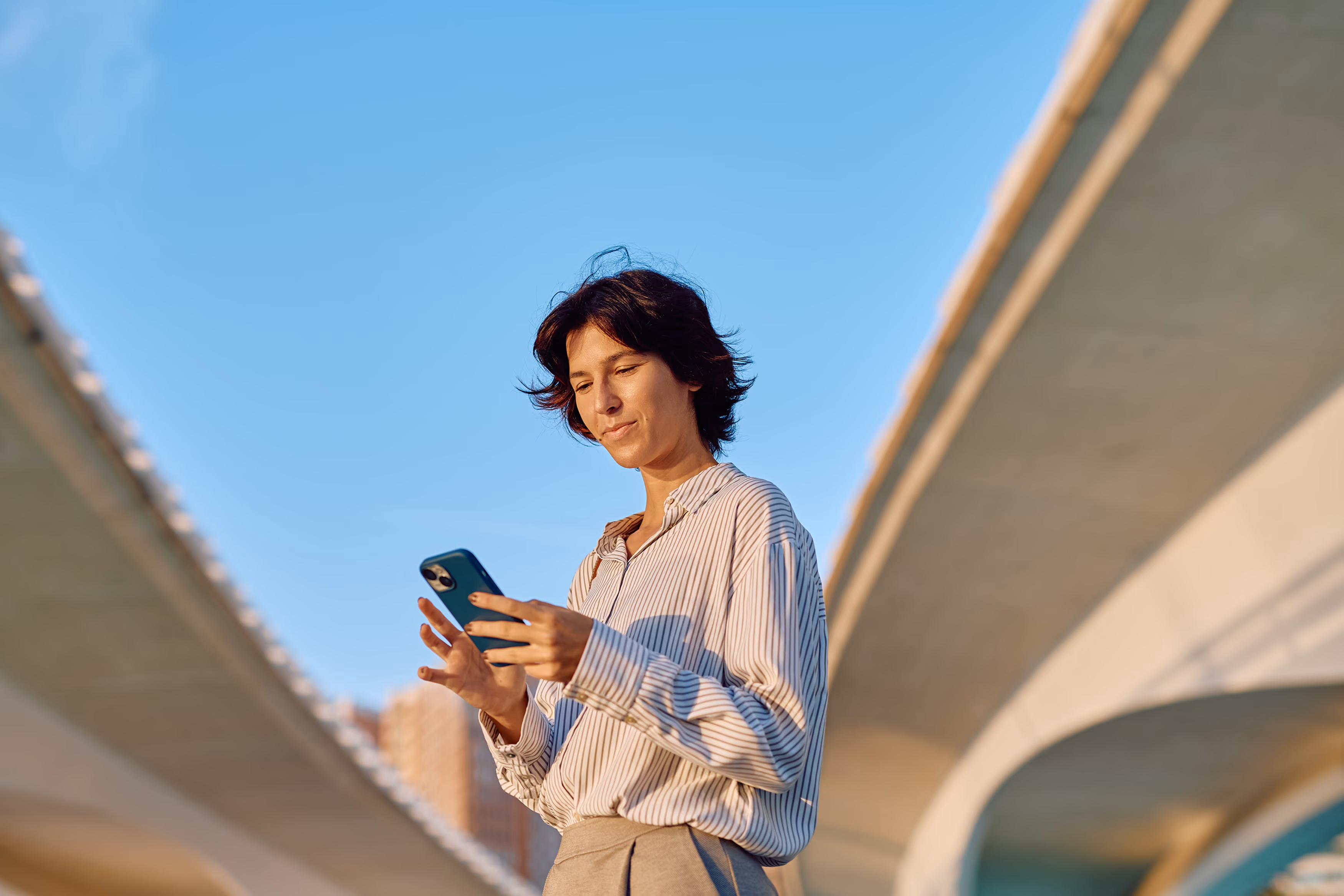 Young person standing outdoors between two overpasses using a smartphone under clear blue sky.