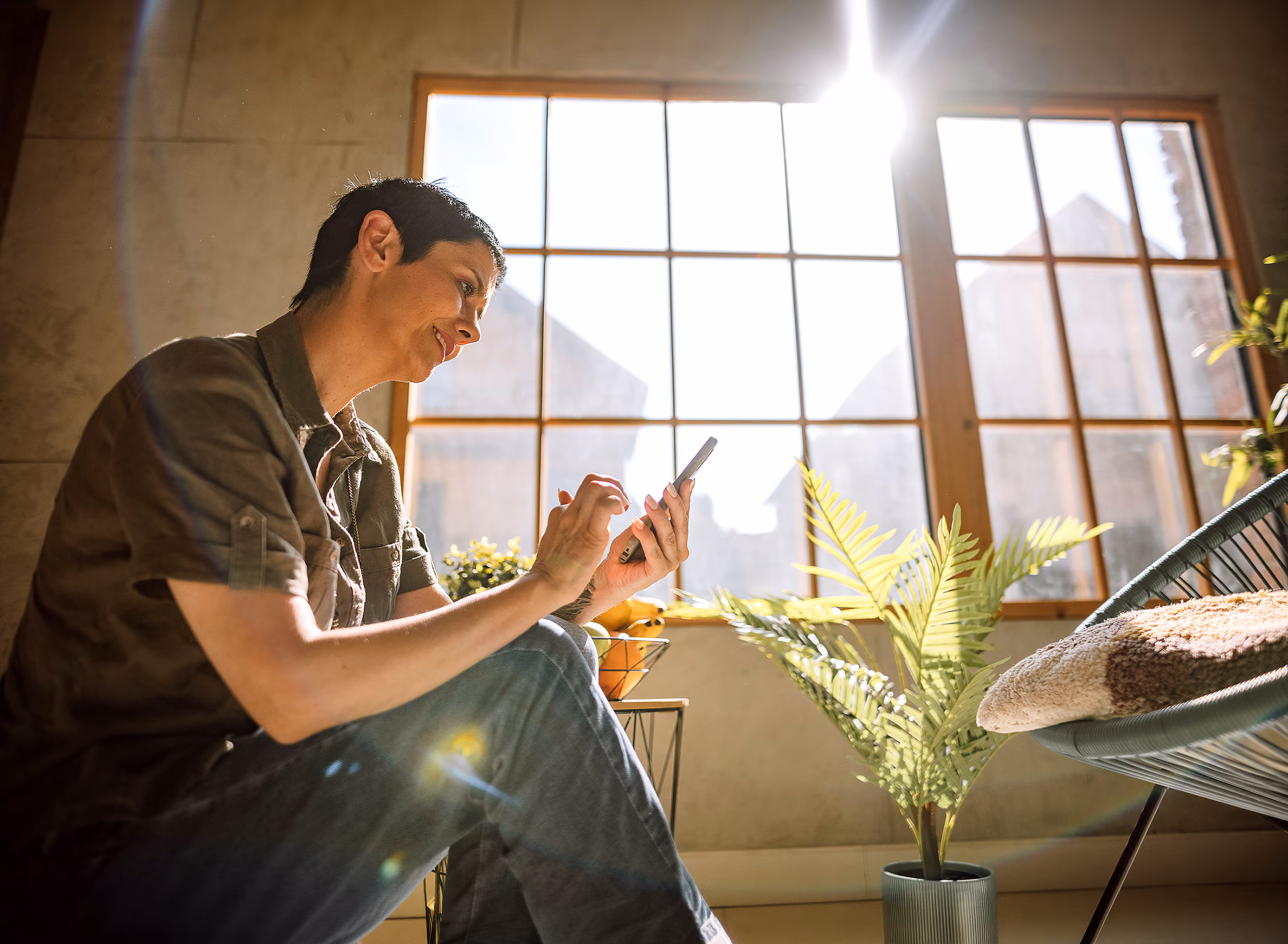 Person sitting indoors by a large window using a smartphone on a sunny day with plants nearby.