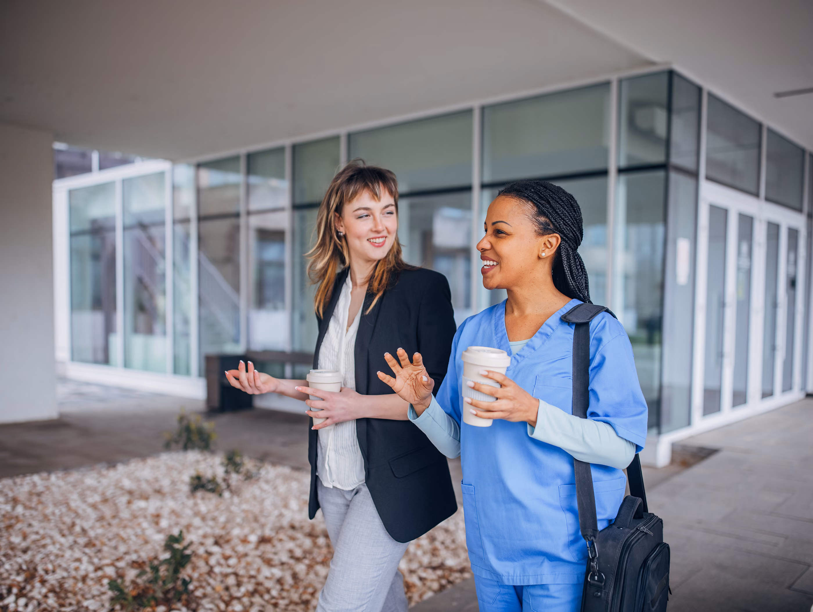 Two women smiling and talking while holding coffee cups outside a modern building, one in scrubs.