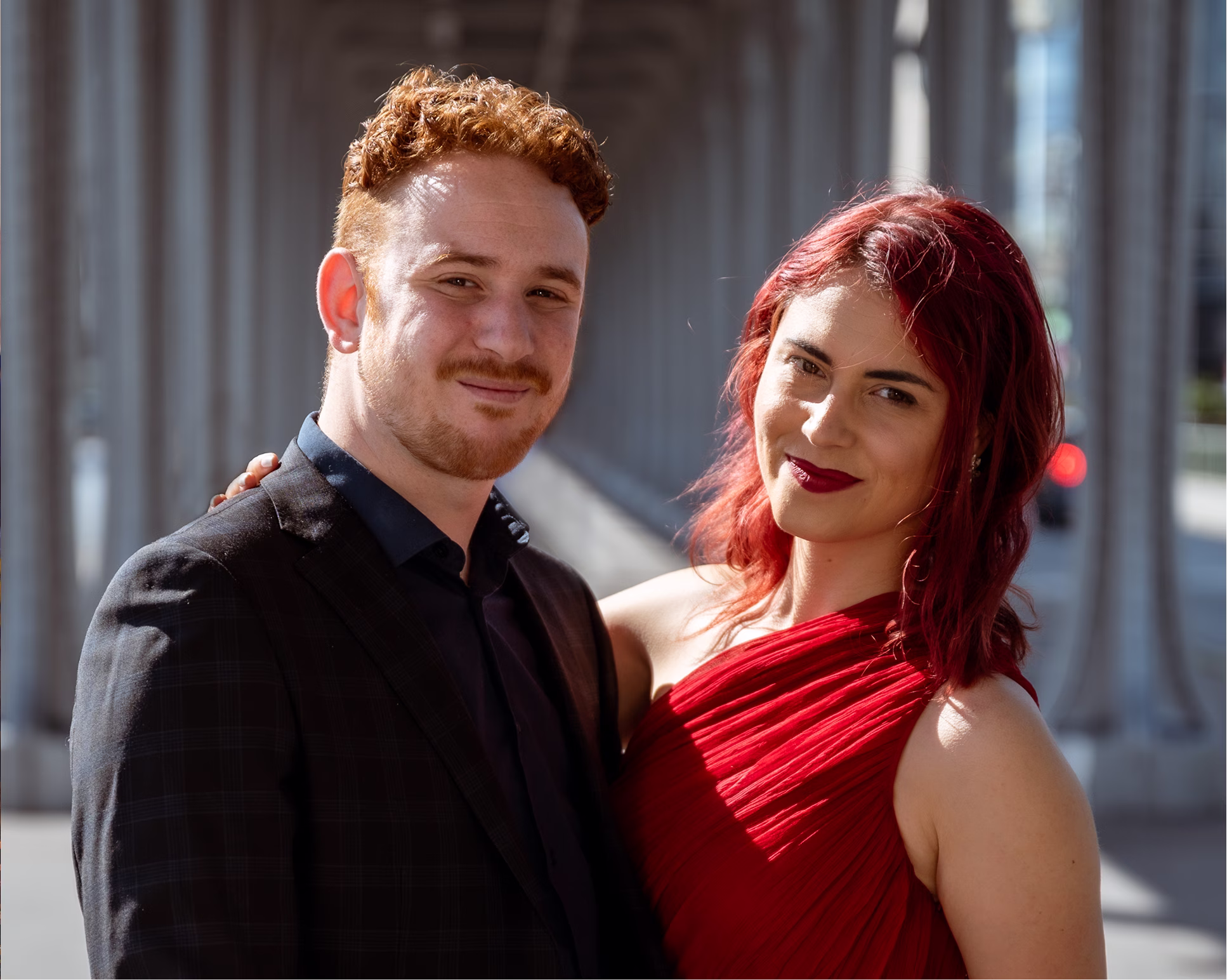 Couple souriant, homme en costume sombre, femme en robe rouge, posant sous une structure en béton.