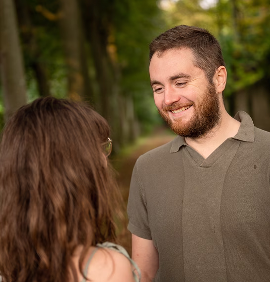 Un homme souriant regarde une femme aux cheveux longs dans un sentier boisé verdoyant.